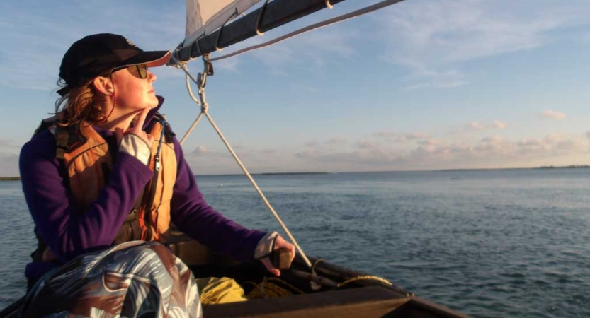 A person wearing a life jacket sits in a sailboat and stares out over the calm blue water. 